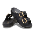 The Crocs Womens Saturday Metallic Buckle Sandals (213316-001) in black feature two wide straps per shoe with large gold metallic buckles, textured footbeds, and slightly elevated heels for added comfort and style.