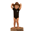 A young child in a Ringers Western Infants S/S Romper (523001RW) Black stands barefoot on a tree stump, holding a large brown cowboy hat above their head, set against a plain white background.