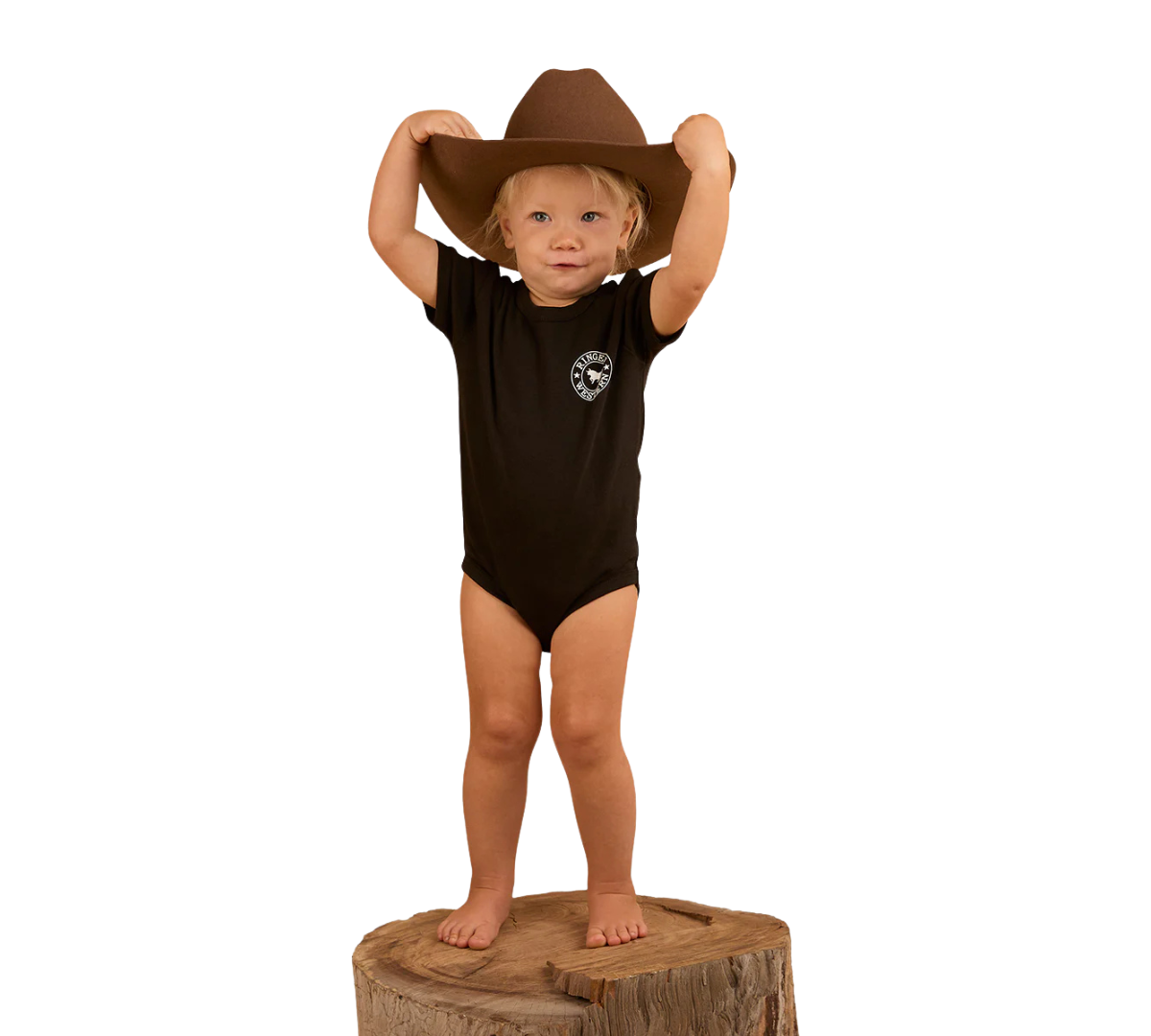 A young child in a Ringers Western Infants S/S Romper (523001RW) Black stands barefoot on a tree stump, holding a large brown cowboy hat above their head, set against a plain white background.