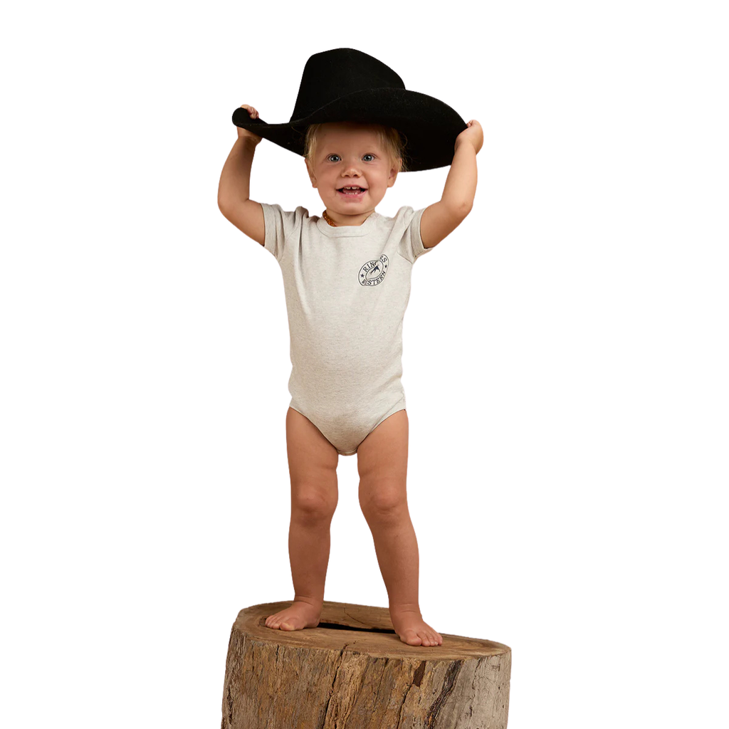 A smiling toddler stands barefoot on a tree stump, wearing the Ringers Western Infants S/S Romper (523001RW) in Grey Marle and holding a large black cowboy hat on their head against a white background.