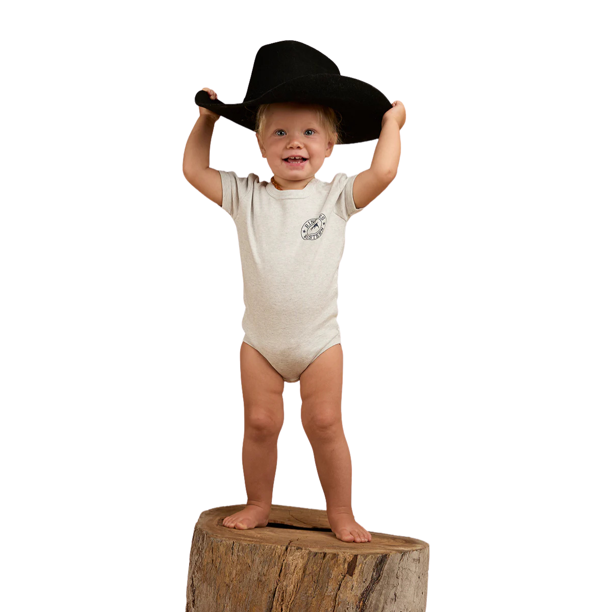 A smiling toddler stands barefoot on a tree stump, wearing the Ringers Western Infants S/S Romper (523001RW) in Grey Marle and holding a large black cowboy hat on their head against a white background.
