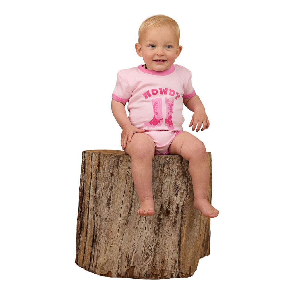 A happy baby girl sits on a tree stump in the Ringers Western Infants Howdy Cowgirl S/S Romper (Ballet Pink), featuring cute cowboy boots print—showcasing the comfort and style of this Ringers Western cotton romper.