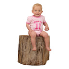 A happy baby girl sits on a tree stump in the Ringers Western Infants Howdy Cowgirl S/S Romper (Ballet Pink), featuring cute cowboy boots print—showcasing the comfort and style of this Ringers Western cotton romper.