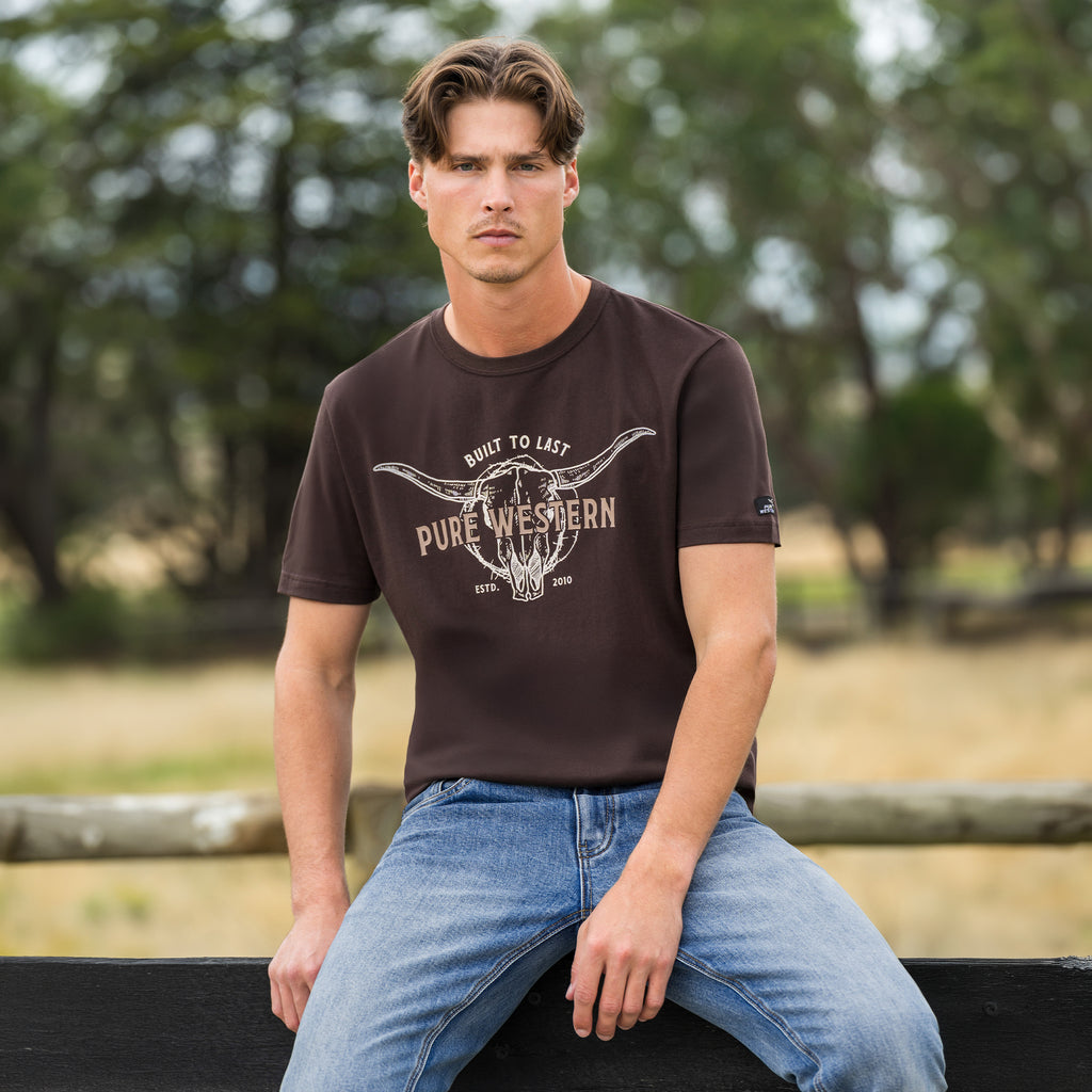 A man wearing a Pure Western Men's Tony Short Sleeve Tee (P6S1503313) in a Chocolate colour with denim jeans sitting on a fence in the bush.