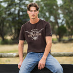 A man wearing a Pure Western Men's Tony Short Sleeve Tee (P6S1503313) in a Chocolate colour with denim jeans sitting on a fence in the bush.
