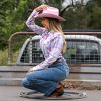 A woman crouching on the back of a ute wearing a Pure Western Storm Hat (PCP2911HAT) in Pink, a check shirt, denim jeans, a western belt and western boots with a western lariat at her feet.
