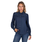 A woman confidently poses against a white background wearing the Pure Western Womens Daniela L/S Shirt (P6W2598204) in blue, paired with blue jeans, one hand touching her shirt.