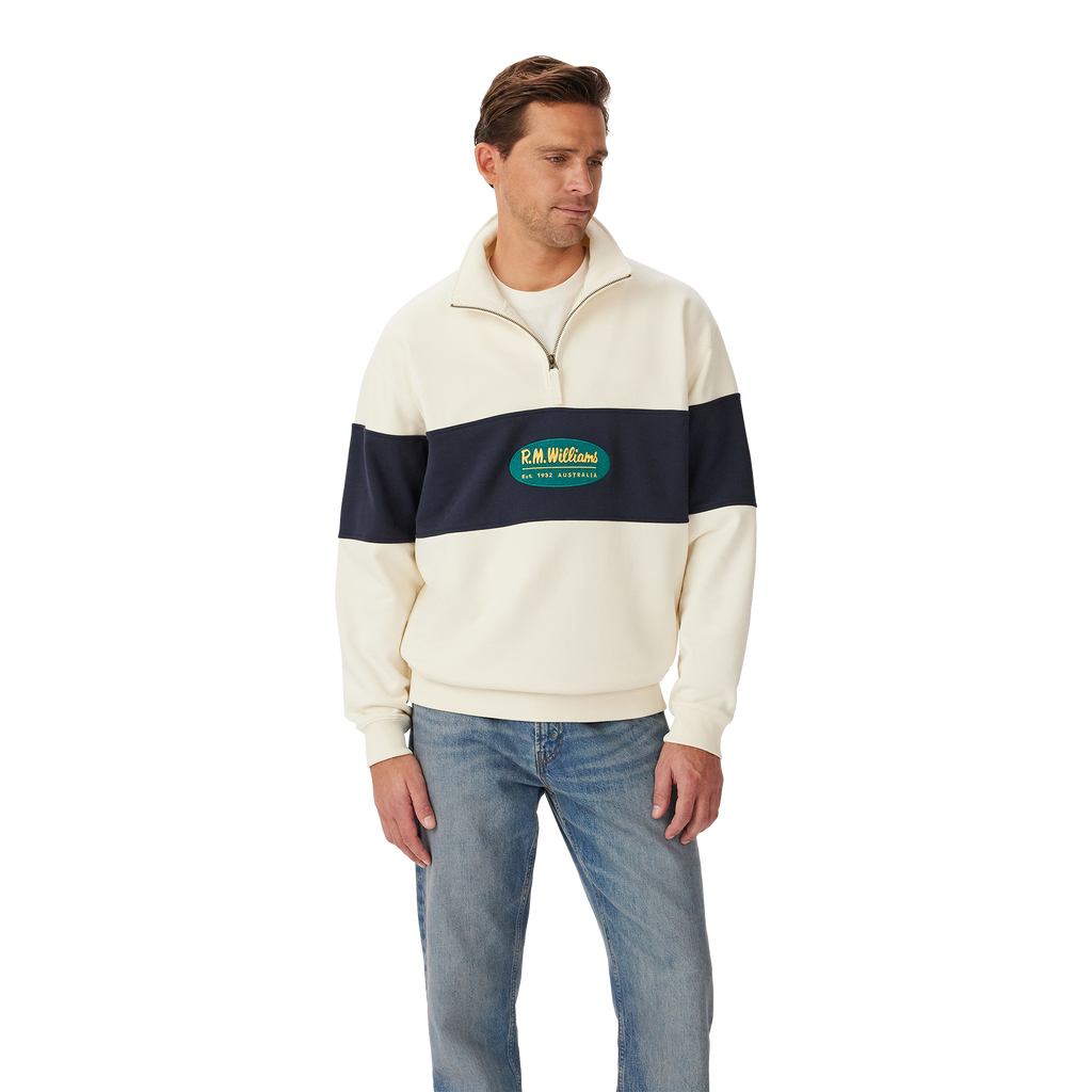 A man stands against a white background wearing the R.M.Williams Men's 90s Logo 1/4 Zip Jumper in ecru with a navy chest and sleeve stripe, paired with light blue jeans.