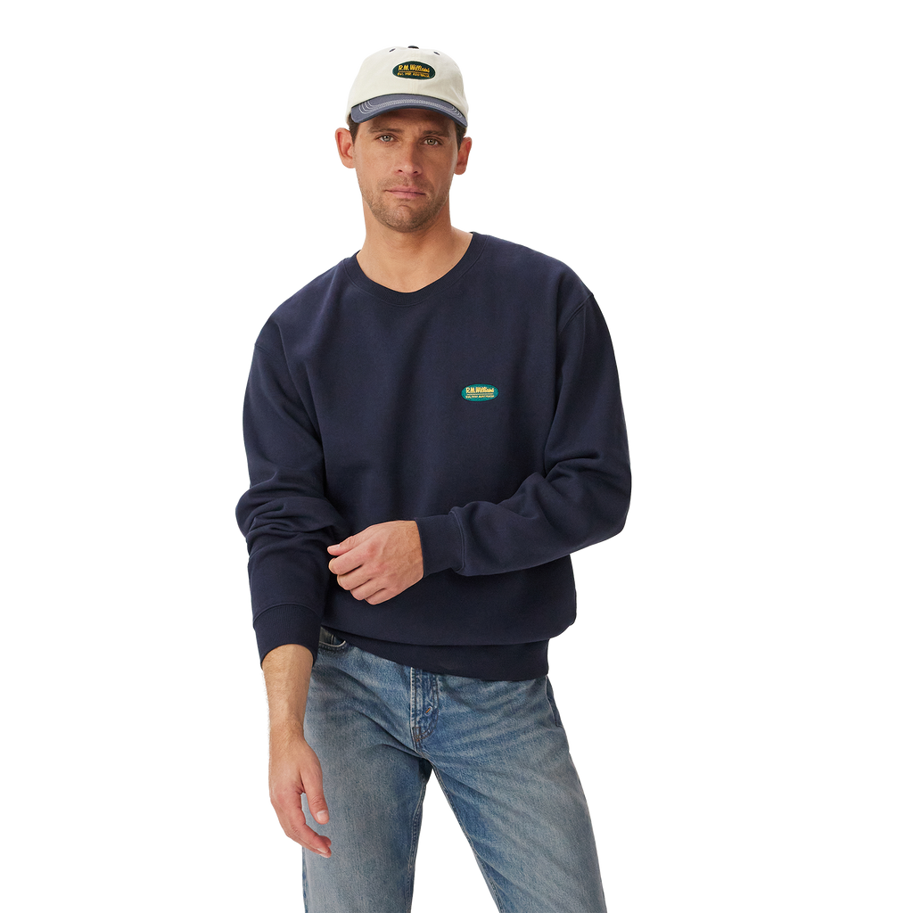 A man stands against a plain white background wearing a vintage navy R.M.Williams Mens 90s Logo Crew Jumper (KCU00BB01), light blue jeans, and a light gray cap.