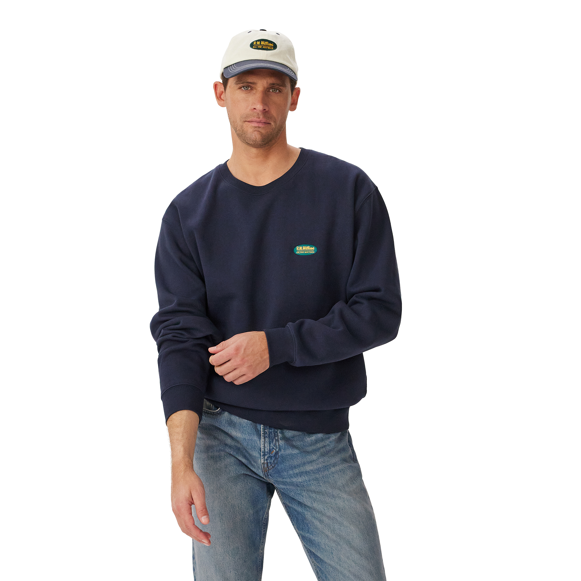A man stands against a plain white background wearing a vintage navy R.M.Williams Mens 90s Logo Crew Jumper (KCU00BB01), light blue jeans, and a light gray cap.