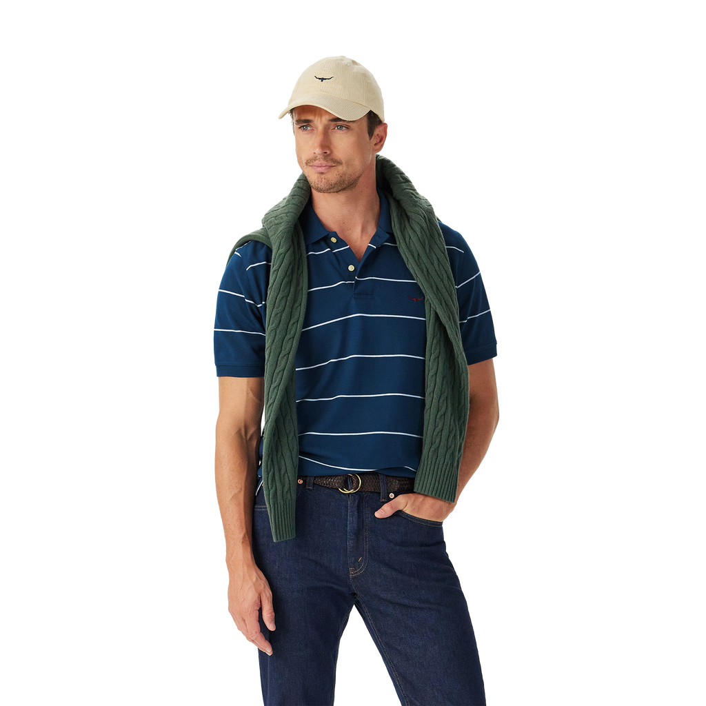 A man in a beige cap, dark jeans, and green sweater draped over his shoulders stands against a white background wearing the R.M.Williams Mens Albany Stripe Polo (KP210QS10) in Vintage Blue for a classic, relaxed look.