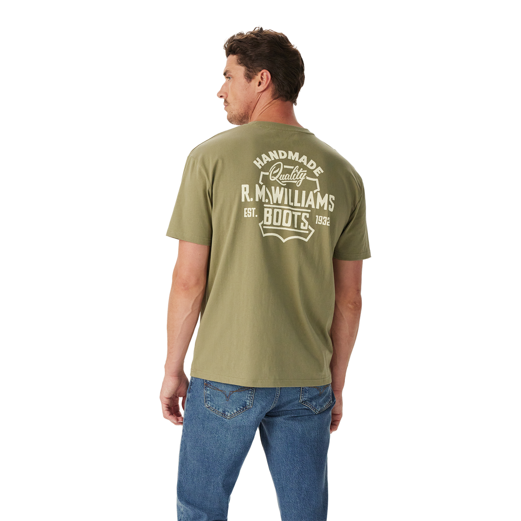 A man wears relaxed fit blue jeans and the R.M.Williams Unisex Boots S/S Tee (KDU10JE08) in Sage, featuring "Handmade Quality R.M.Williams Boots Est. 1932" printed in white on the back, standing against a white background.