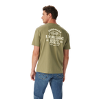 A man wears relaxed fit blue jeans and the R.M.Williams Unisex Boots S/S Tee (KDU10JE08) in Sage, featuring "Handmade Quality R.M.Williams Boots Est. 1932" printed in white on the back, standing against a white background.