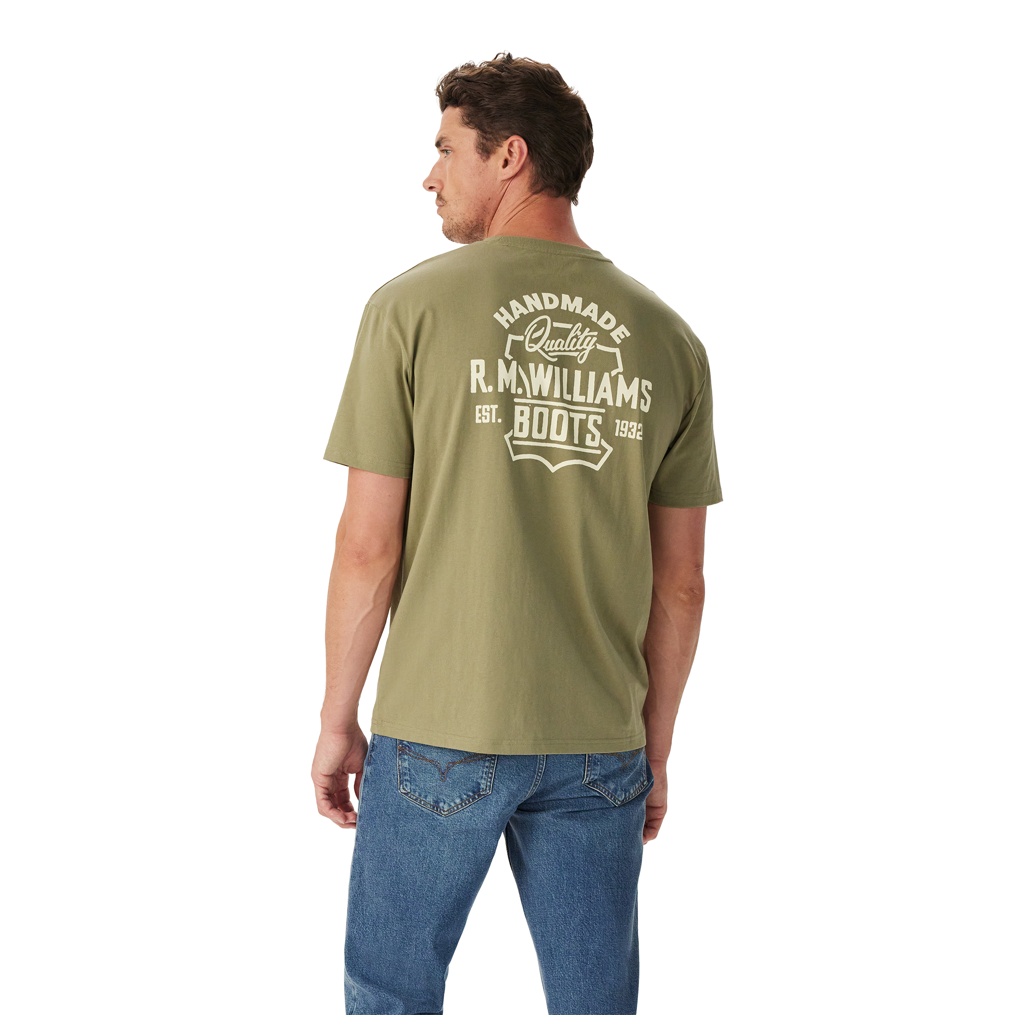 A man wears relaxed fit blue jeans and the R.M.Williams Unisex Boots S/S Tee (KDU10JE08) in Sage, featuring "Handmade Quality R.M.Williams Boots Est. 1932" printed in white on the back, standing against a white background.