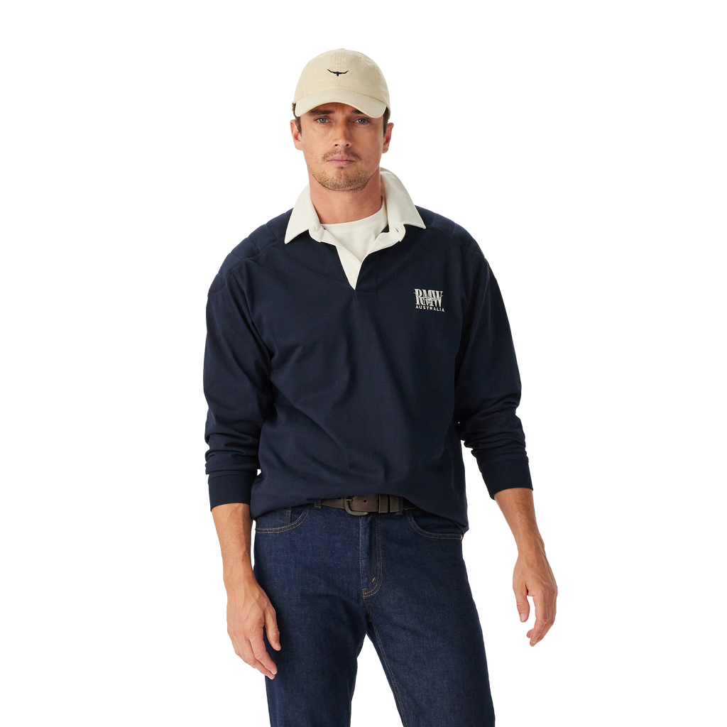 Man wearing a navy R.M.Williams Classic Shoulder Rugby with an R.M.Williams logo, beige cap, brown leather belt, and dark jeans on a white background. R.M.Williams Mens Classic Shoulder Rugby (KR200RJ4502) Navy