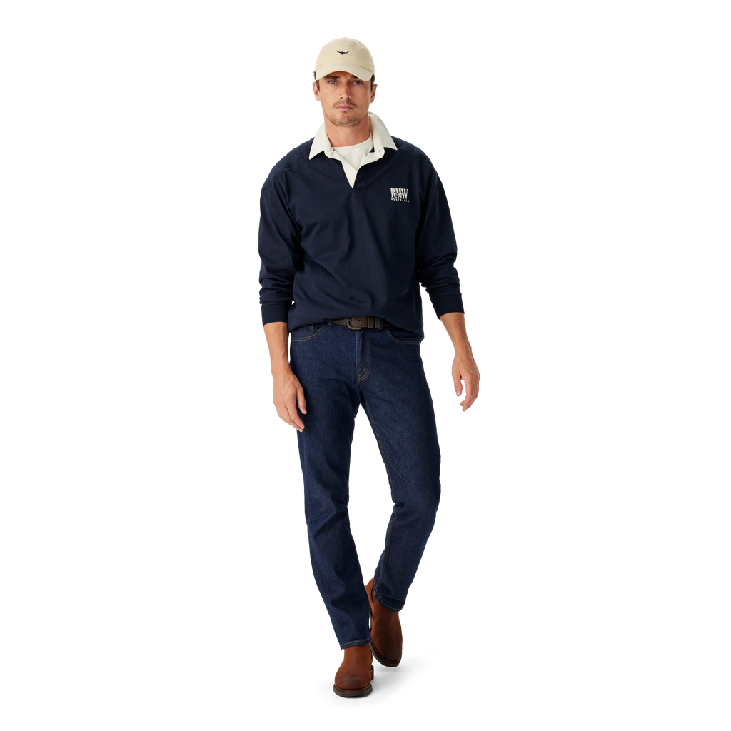 Man wearing a navy R.M.Williams Classic Shoulder Rugby with an R.M.Williams logo, beige cap, brown leather belt, brown boots and dark jeans on a white background.  R.M.Williams Mens Classic Shoulder Rugby (KR200RJ4502) Navy