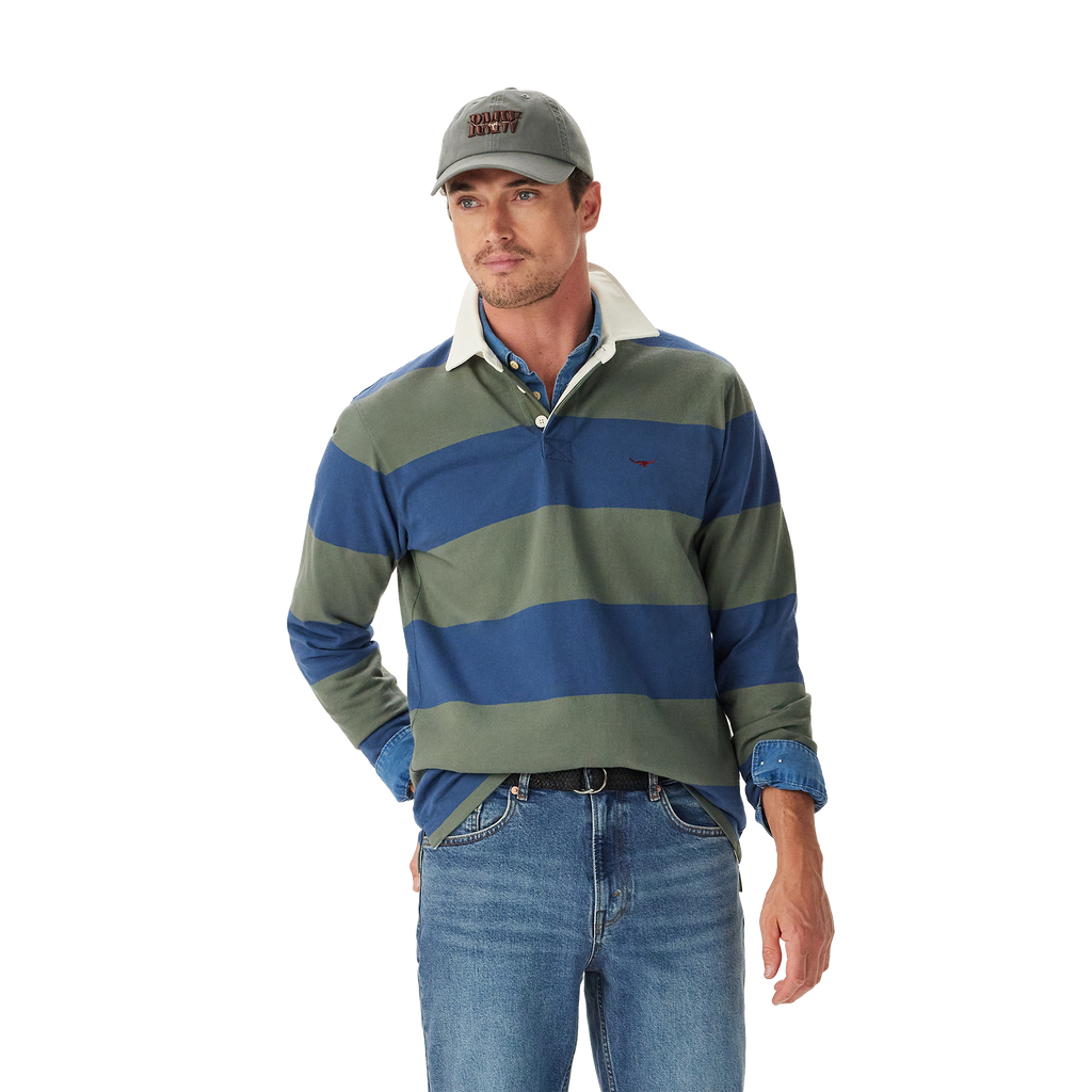 A man wearing an R.M.Williams Mens Jones Stripe Rugby (KR200RJ16) in Ensign Blue, a gray cap, and blue jeans stands with one hand in his pocket against a plain white background.