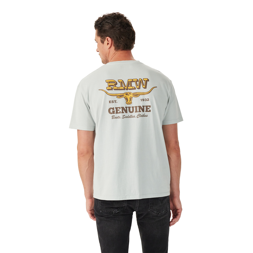 A man in black jeans faces away, wearing the R.M.Williams Mens Louth S/S Tee (KDU10JE05) Salt—a light grey, relaxed fit tee with a yellow graphic and "RMW EST. 1932 GENUINE Boots, Saddles, Clothes" above a longhorn skull on the back.