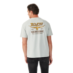A man in black jeans faces away, wearing the R.M.Williams Mens Louth S/S Tee (KDU10JE05) Salt—a light grey, relaxed fit tee with a yellow graphic and "RMW EST. 1932 GENUINE Boots, Saddles, Clothes" above a longhorn skull on the back.