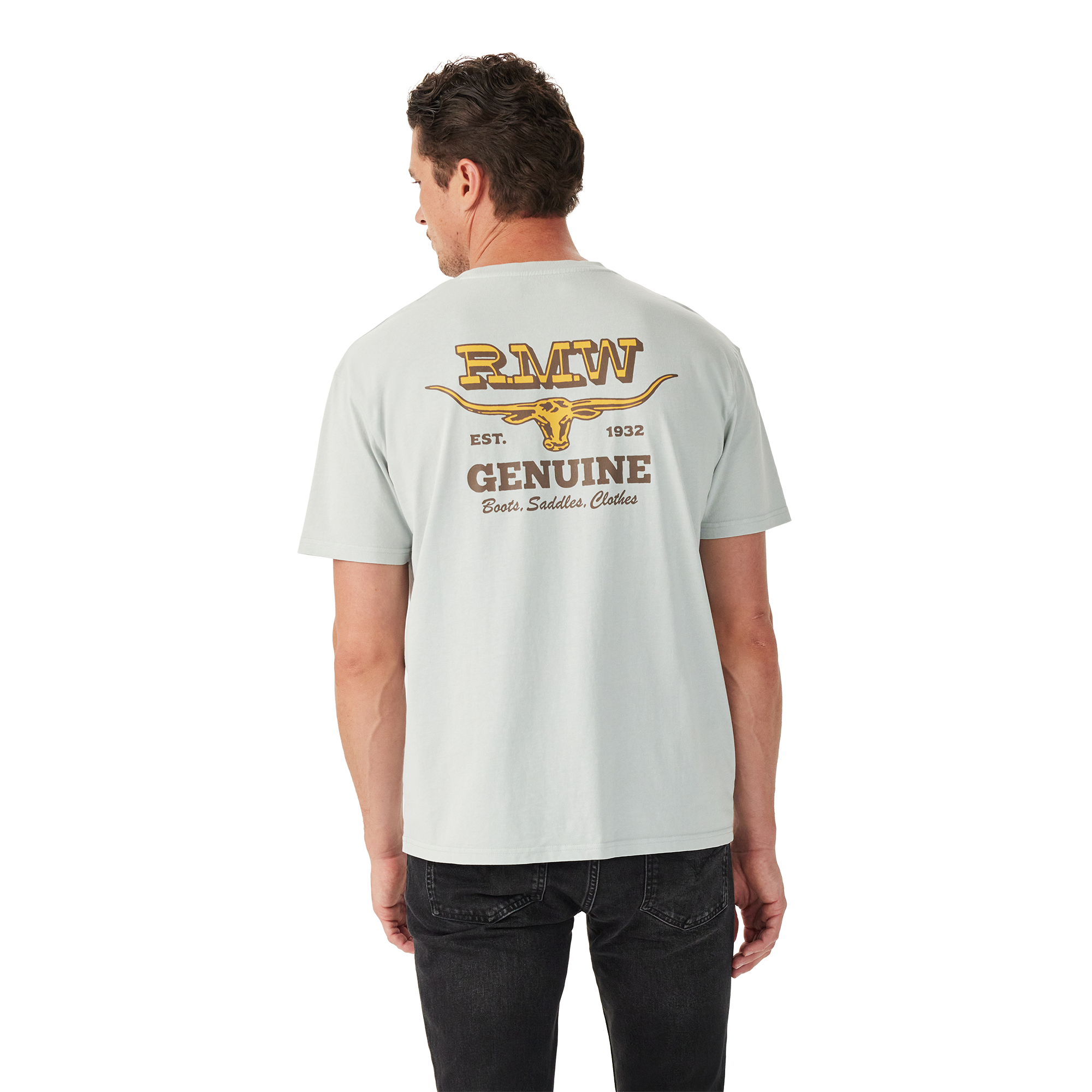 A man in black jeans faces away, wearing the R.M.Williams Mens Louth S/S Tee (KDU10JE05) Salt—a light grey, relaxed fit tee with a yellow graphic and "RMW EST. 1932 GENUINE Boots, Saddles, Clothes" above a longhorn skull on the back.