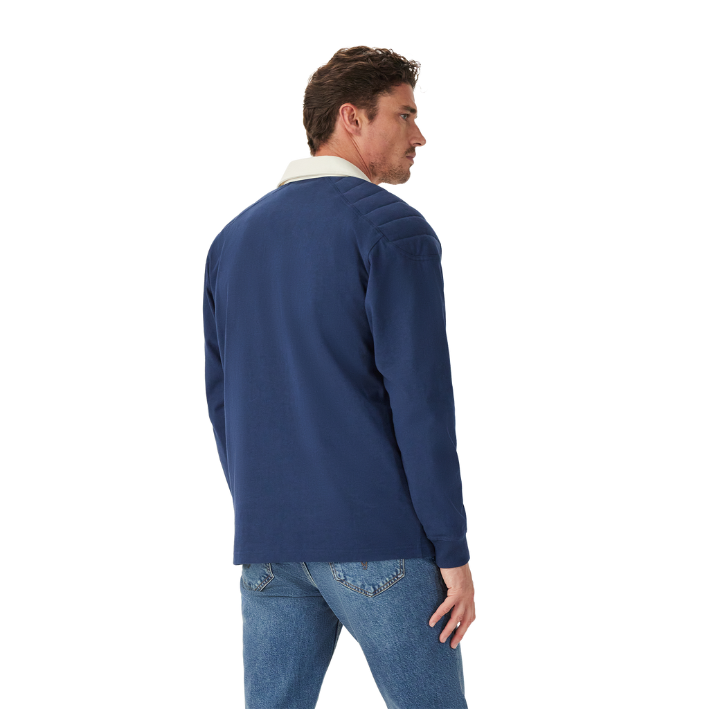 A man stands casually with his back partially to the camera, wearing the R.M.Williams Mens Mulyungarie 1/4 Zip Sweatshirt (KZU00BB03) in Vintage Blue over a light blue shirt and blue jeans, against a plain white background.