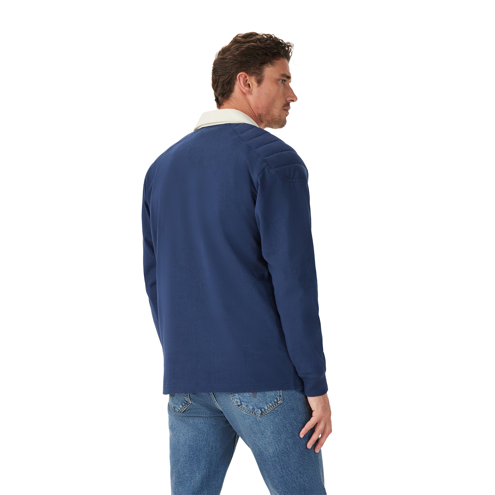 A man stands casually with his back partially to the camera, wearing the R.M.Williams Mens Mulyungarie 1/4 Zip Sweatshirt (KZU00BB03) in Vintage Blue over a light blue shirt and blue jeans, against a plain white background.