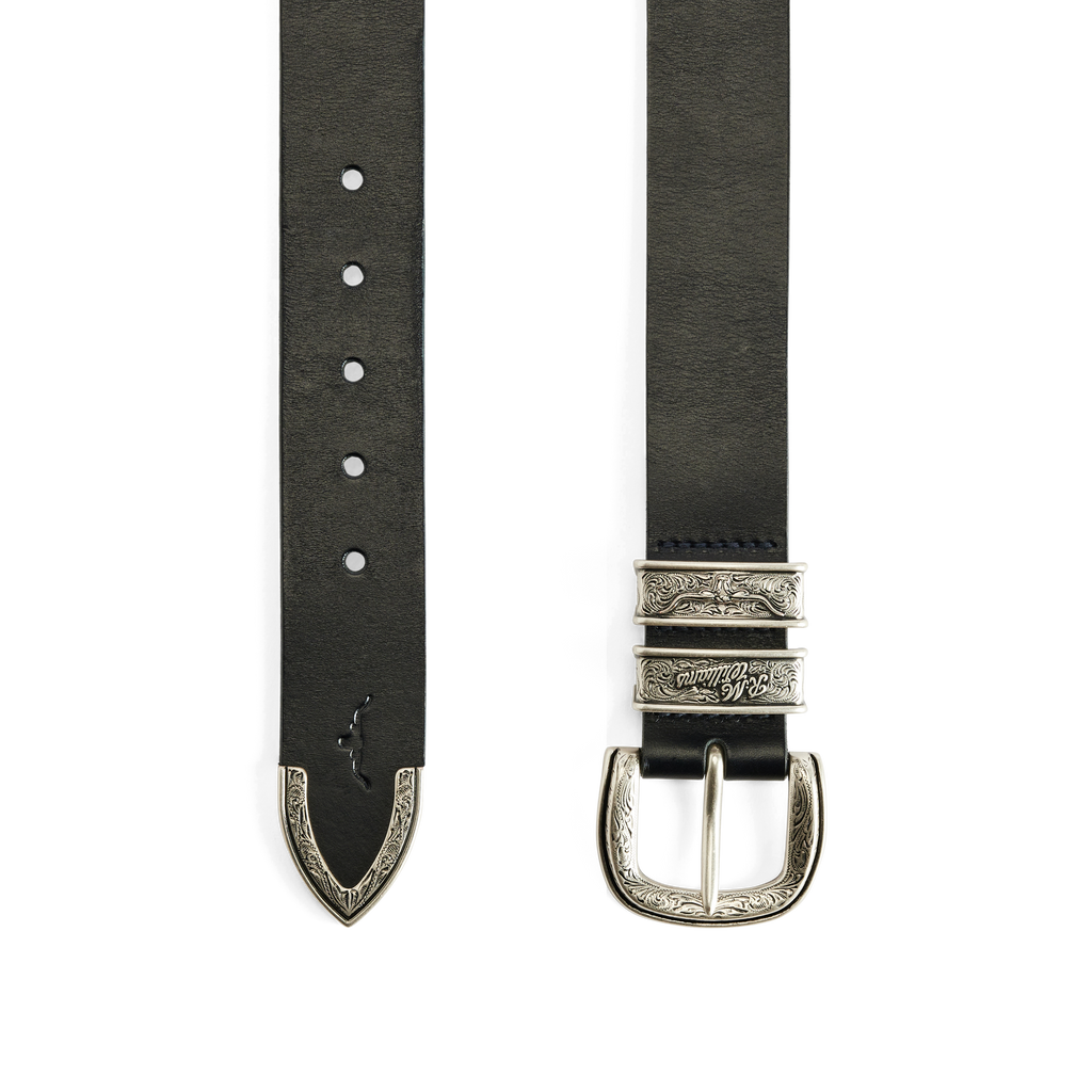 Flat lay of the engraved and embossed silver toned buckle, keepers and tail of a black R.M.Williams Western Drover Belt (BSF5DCH02) on a white background.