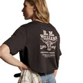A woman from behind wearing a dark chocolate R.M.Williams Kandos Short Sleeve Tee (KDX10JE06) with a large back RMW longhorn logo print, paired with denim jeans and a plaited leather belt, with her hand on her hip, which is holding a check shirt, photographed on a white background.