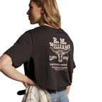 A woman from behind wearing a dark chocolate R.M.Williams Kandos Short Sleeve Tee (KDX10JE06) with a large back RMW longhorn logo print, paired with denim jeans and a plaited leather belt, with her hand on her hip, which is holding a check shirt, photographed on a white background.