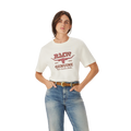 A woman wears the R.M.Williams Womens Louth S/S Tee (KDR10JE37) in white with red "RAW GENUINE" text, paired with blue Australian cotton jeans and a black belt, standing hands-in-pockets against a plain white background.