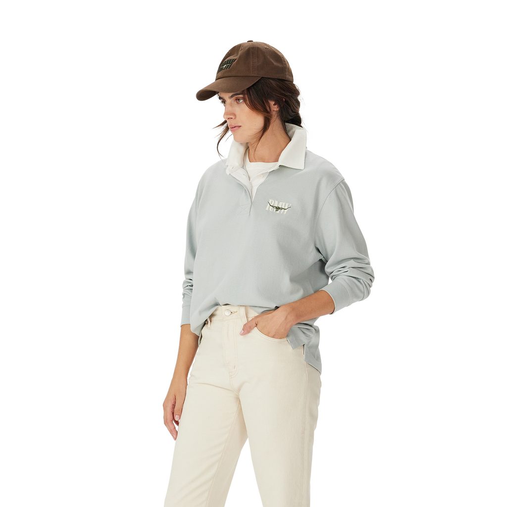 A woman in the R.M.Williams Womens Narran Heritage Rugby (Salt), featuring a light gray collared organic cotton design with Heritage logo embroidery, brown cap, and off-white pants stands with one hand in her pocket against a plain white background.