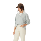 A woman in the R.M.Williams Womens Narran Heritage Rugby (Salt), featuring a light gray collared organic cotton design with Heritage logo embroidery, brown cap, and off-white pants stands with one hand in her pocket against a plain white background.