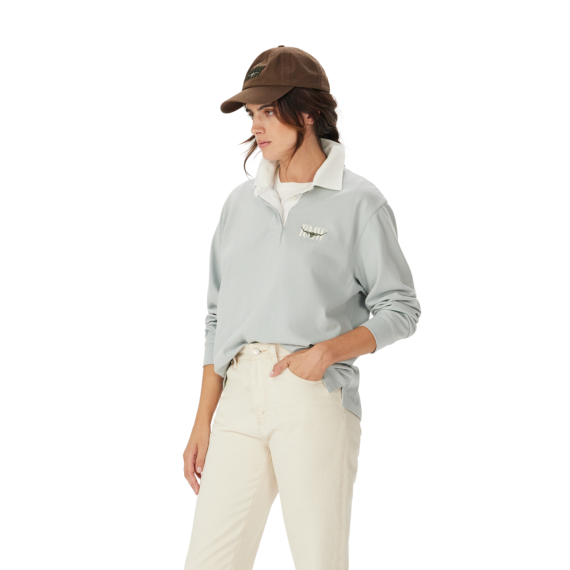 A woman in the R.M.Williams Womens Narran Heritage Rugby (Salt), featuring a light gray collared organic cotton design with Heritage logo embroidery, brown cap, and off-white pants stands with one hand in her pocket against a plain white background.