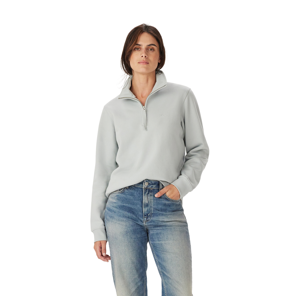 A woman with straight brown hair and a neutral expression wears the R.M.Williams Womens Trickett 1/4 Zip Sweatshirt (Salt) and blue jeans, standing against a plain white background with one hand in her pocket.