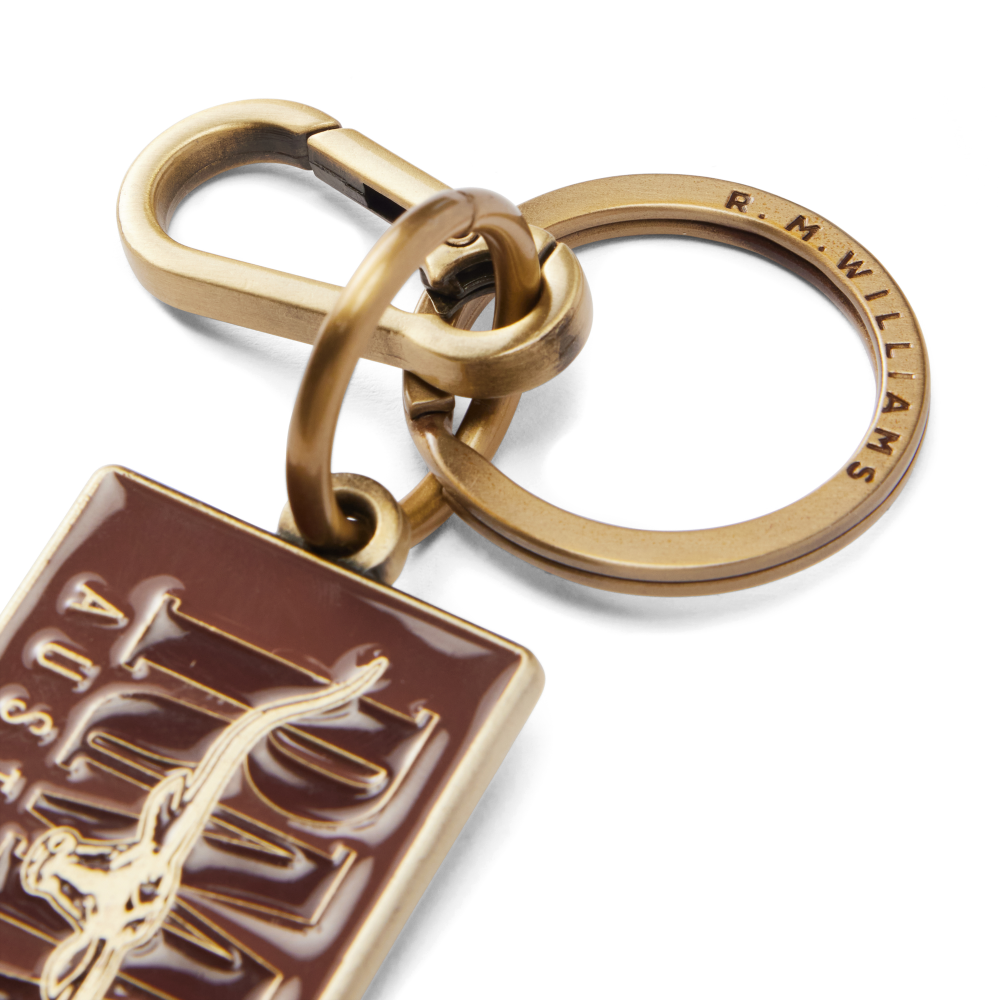 A close up of an R.M.Williams Men's Heritage Logo Enamel Keyring (CKS4AMT01) with a brass clasp and a brown enamel tag featuring 'R.M. Williams Australia' on a white background.