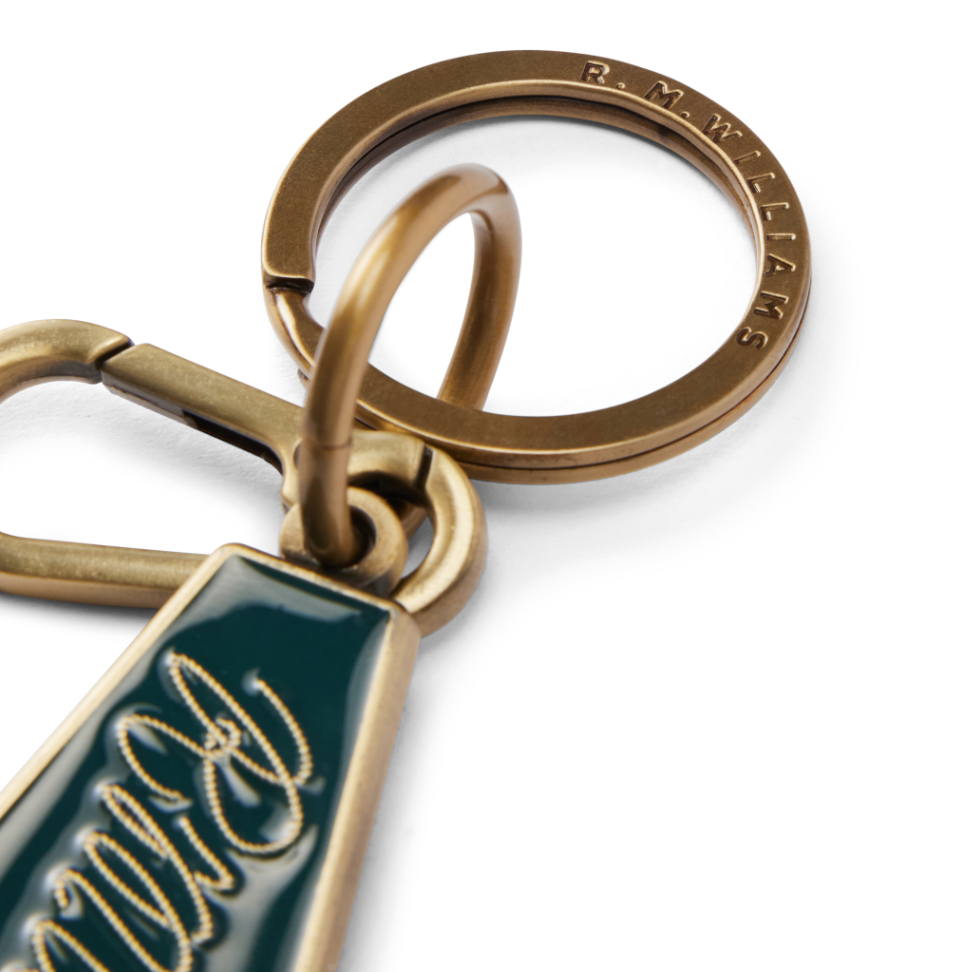 Close up detail of a brass and pine green R.M.Williams Men's Rope Logo Enamel Keyring (CKS4AMT01) with cursive RMWilliams text on a white background.