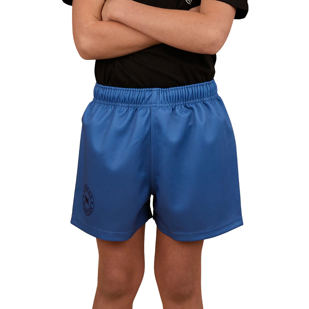 A child in a black shirt and Ringers Western Childrens Big Ringers Footy Shorts (325020RW) Blue stands arms crossed. The blue shorts, branded by Ringers Western, have an elastic waistband, left-leg emblem, and durable design. Face not visible.
