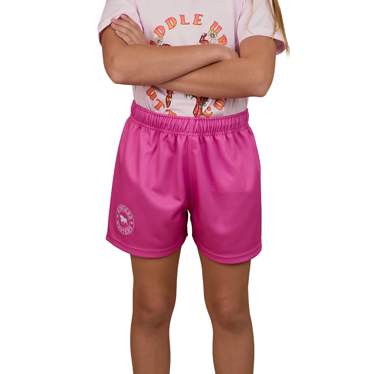 A child with arms crossed wears a light pink T-shirt and Ringers Western Childrens Big Ringers Footy Shorts (Candy), featuring the brand's emblem on the left leg. The child's face is not visible.
