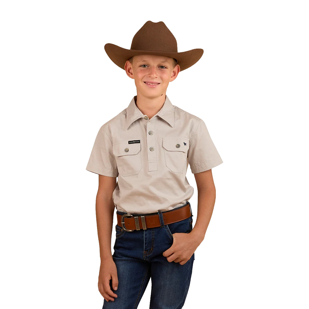 A young boy wears the Ringers Western Childrens Ord Half Button S/S Work Shirt in beige, paired with a brown cowboy hat, blue jeans, and a brown belt. He stands smiling with one hand in his pocket against a white background.