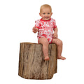 A smiling baby in a barely pink Ringers Western Infants Boot Scootin S/S Romper (524005RW) sits on a large tree stump against a white background.