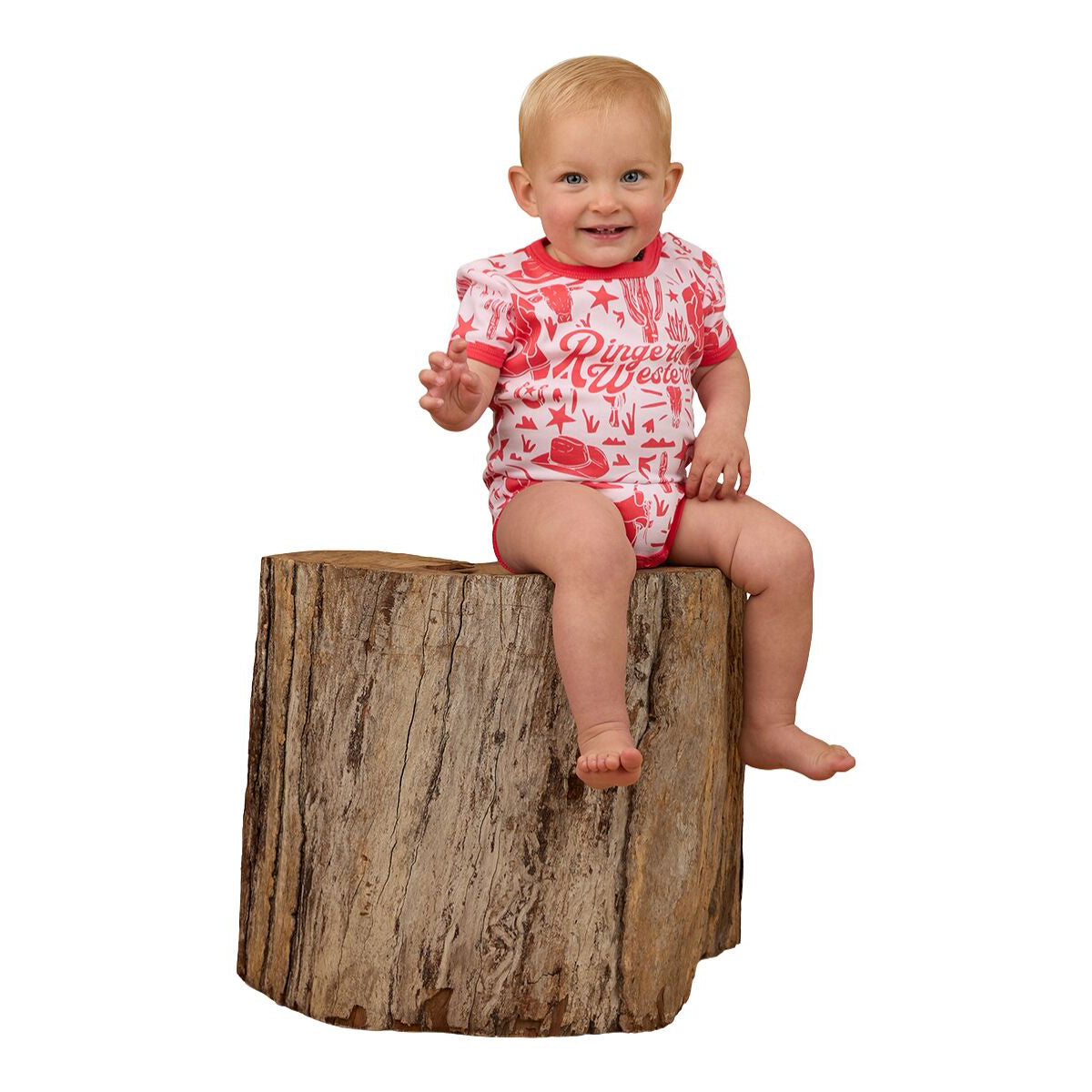 A smiling baby in a barely pink Ringers Western Infants Boot Scootin S/S Romper (524005RW) sits on a large tree stump against a white background.