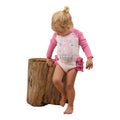 A young blonde girl in a ponytail, barefoot, stands by a large tree stump wearing the Ringers Western Infants Rodeo Gal L/S Frill Romper (White/Pink).