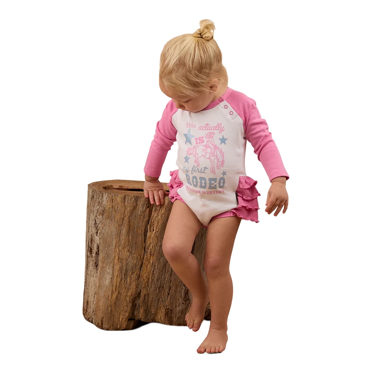 A young blonde girl in a ponytail, barefoot, stands by a large tree stump wearing the Ringers Western Infants Rodeo Gal L/S Frill Romper (White/Pink).