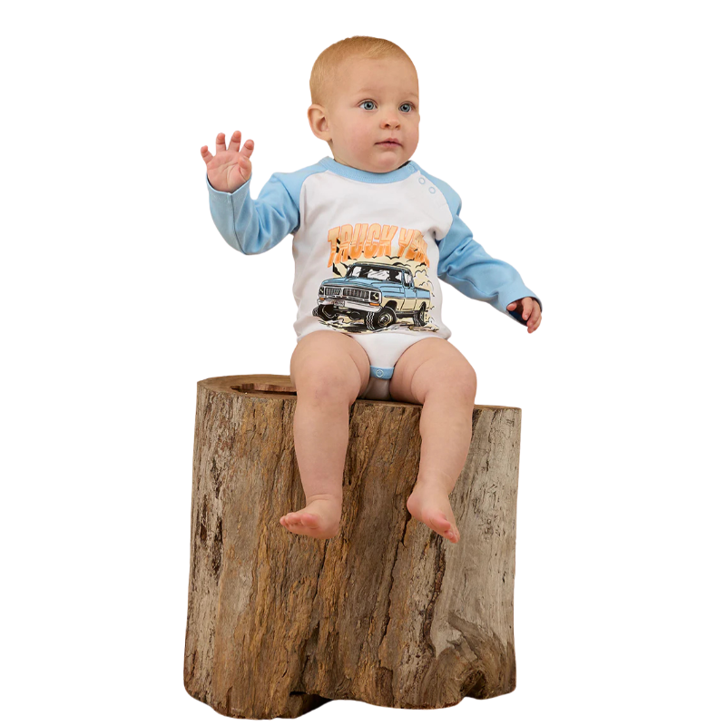 A baby wearing the Ringers Western Infants Trucker Kid L/S Romper in White/Blue sits on a large tree stump with one hand raised, gazing to the side against a plain white background.
