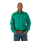 A man wears the Ringers Western Mens King River Half Button Work Shirt in green with blue jeans and a brown belt with a large buckle. He faces forward, smiling with his hands in his pockets, against a plain white background.