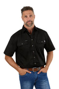 A man with short gray hair and a beard wears a Ringers Western Mens Lake Argyle S/S Full Button Work Shirt (118110004) in black, blue jeans, and a brown belt, standing with his hands in his pockets against a white background.
