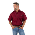 A man with short brown hair and a beard wears the Ringers Western Mens Pack Saddle S/S Half Button Work Shirt (118110003) in Burgundy, paired with blue jeans and a brown belt. He stands against a white background, looking left with hands in his pockets.