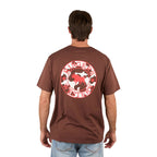 A man stands with his back to the camera in light blue jeans, wearing a loose fit Ringers Western Mens Signature Bull S/S Tee (Chocolate/Red/Cow Print) featuring a large red and white logo and cow graphic on the back.