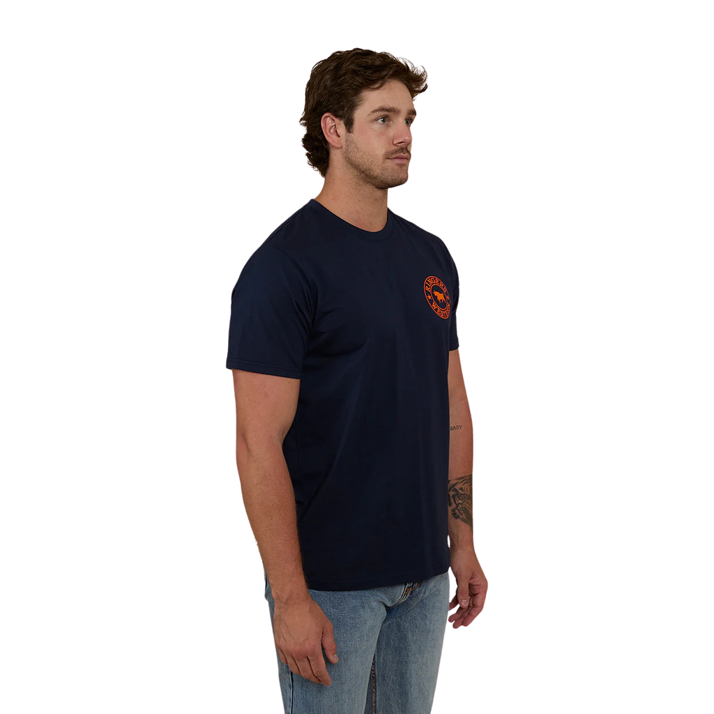 A man wearing a navy blue Ringers Western Men's Signature Bull Loose Fit S/S Tee (120201560) with an orange circular graphic that reads 'Ringers Western' with a bull silhouette, paired with blue jeans against a plain white background.