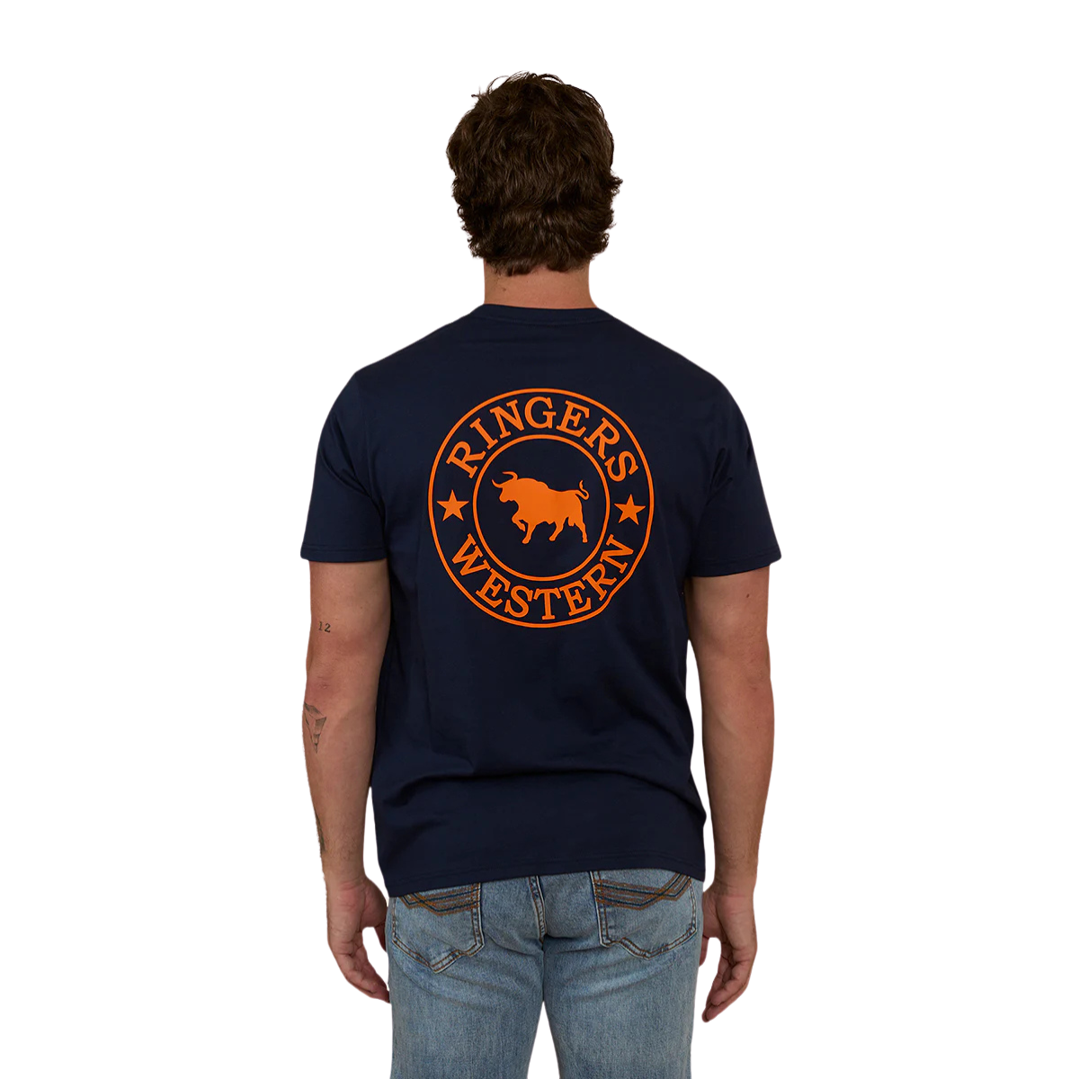 A man viewed from behind wearing a navy blue Ringers Western Men's Signature Bull Loose Fit S/S Tee (120201560) with a large orange circular graphic on the back that reads 'Ringers Western' with a bull silhouette, paired with blue jeans against a plain white background.